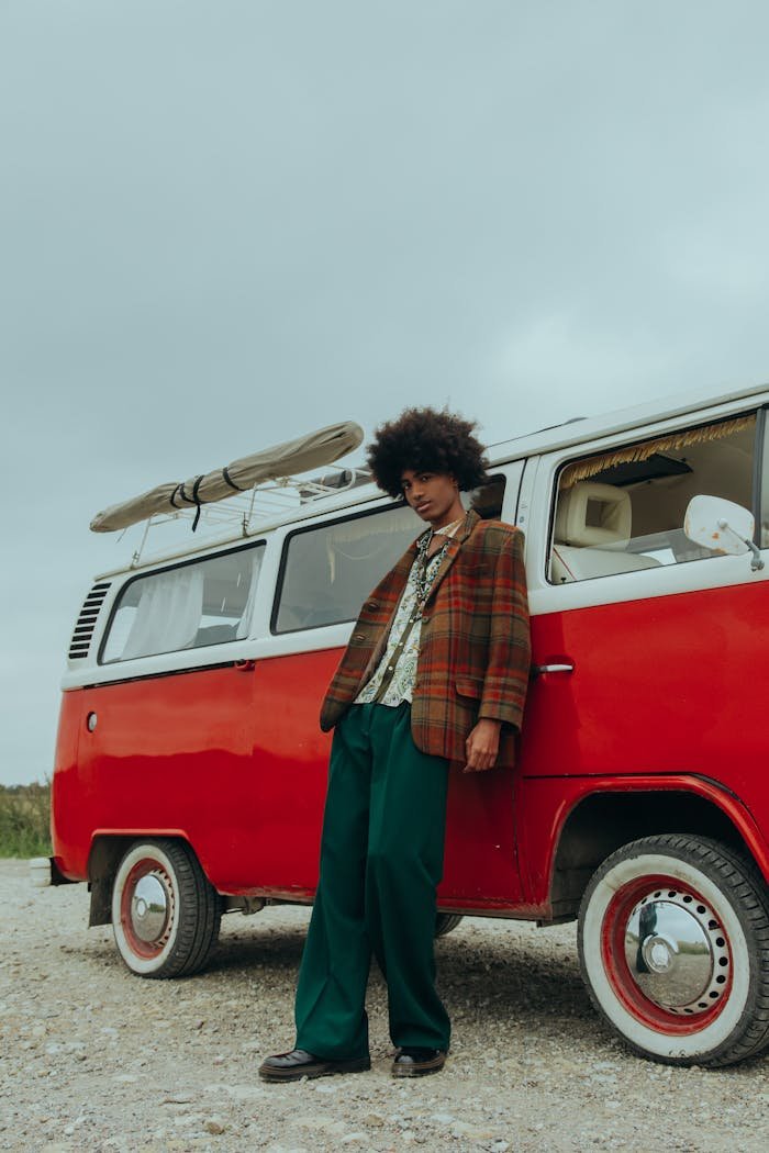 Stylish man with afro leaning on a retro red van outdoors under a cloudy sky.